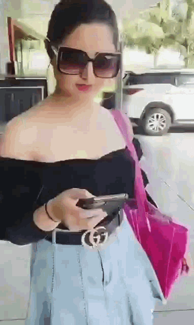 a woman wearing sunglasses and a black off the shoulder top is holding a cell phone .