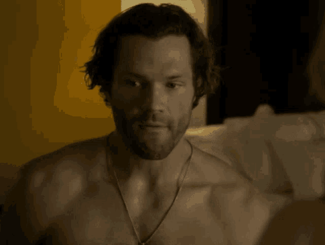 a shirtless man with a necklace around his neck is sitting on a bed