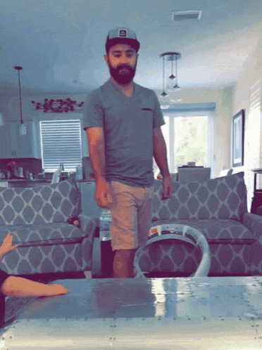 a man with a beard is standing in a living room wearing a hat with the letter e on it
