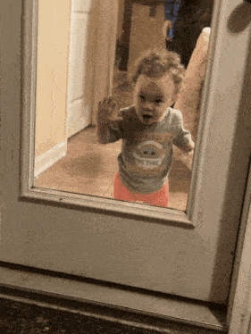 a toddler wearing a shirt that says " the mandalorian " looks through a glass door