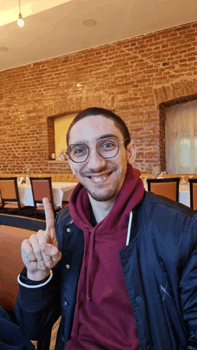 a man wearing glasses and a maroon hoodie is giving the peace sign