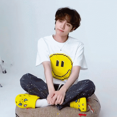 a man wearing a white t-shirt with a pixelated smiley face on it sits on a pillow with his legs crossed