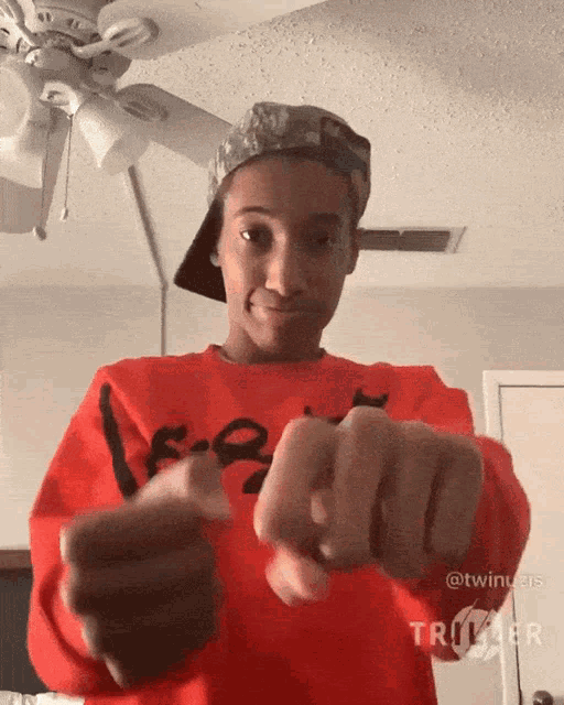 a young man wearing a hat and a red sweater is pointing his finger at the camera