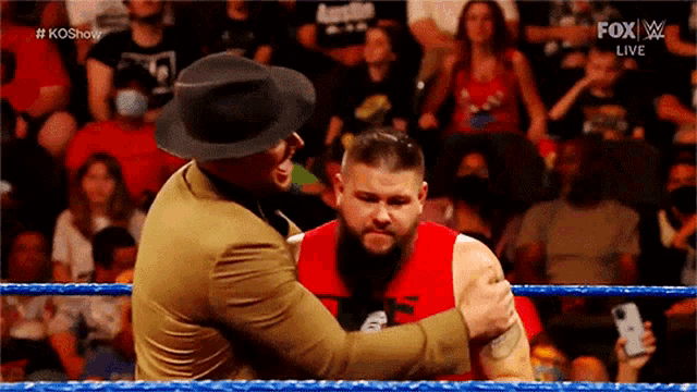 a man in a hat is hugging another man in a red shirt in a wrestling ring