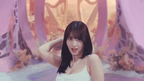 a woman in a white tank top is standing in front of a pink wall and flowers .