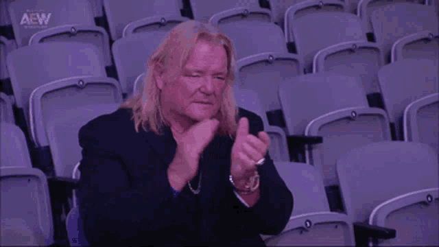 a man with long blonde hair is sitting in an empty stadium and clapping his hands .