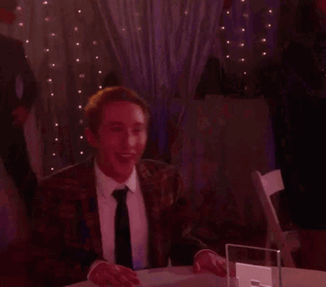 a man in a suit and tie is sitting at a table with purple lights behind him