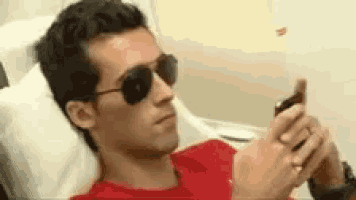 a man wearing sunglasses and a red shirt is laying on a bed looking at his cell phone .