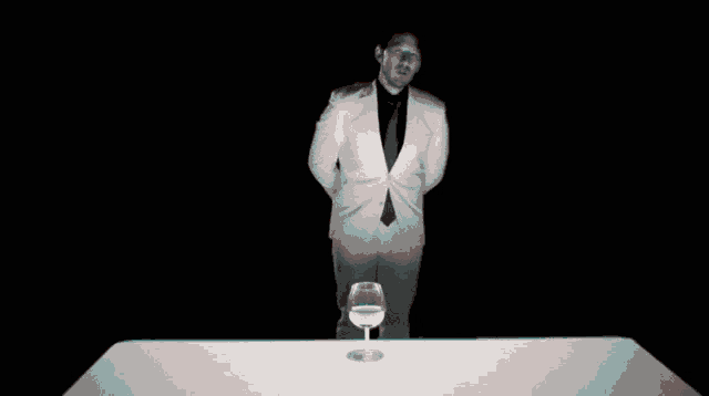 a man in a white suit and black tie stands in front of a glass of water