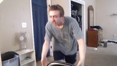 a man in a grey shirt is standing in a room with a fan in the background