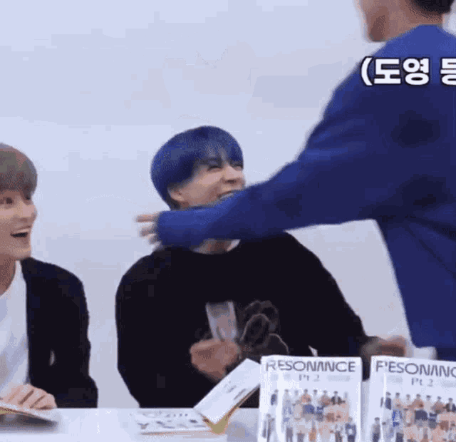 a man with blue hair is hugging another man while sitting at a table with two other men .