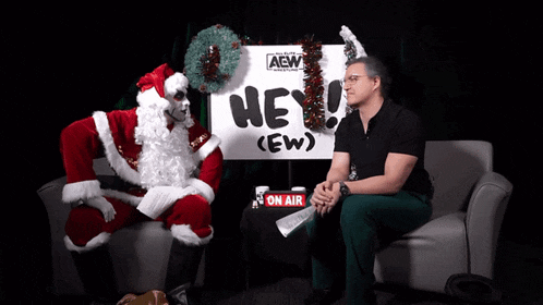 a man in a santa suit sits in front of a sign that says hey ( ew ) on it