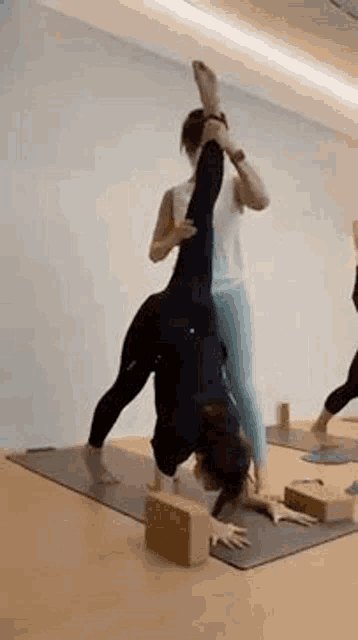 a woman is doing a handstand on a yoga mat while another woman holds her leg over her head .