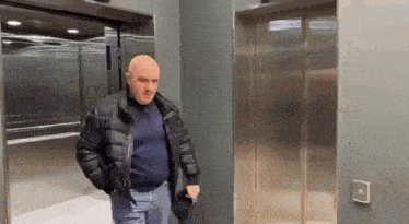 a bald man in a black jacket is standing in an elevator with the doors open .