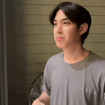 a young man wearing a gray t-shirt that says super sun is standing in front of a window .