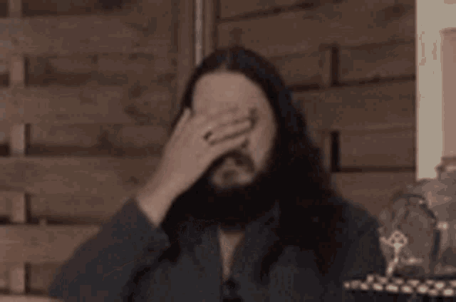 a man with long hair and a beard is covering his face with his hand while sitting at a table .