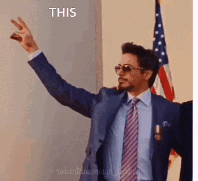 robert downey jr. is wearing a suit and tie and giving a peace sign in front of an american flag .