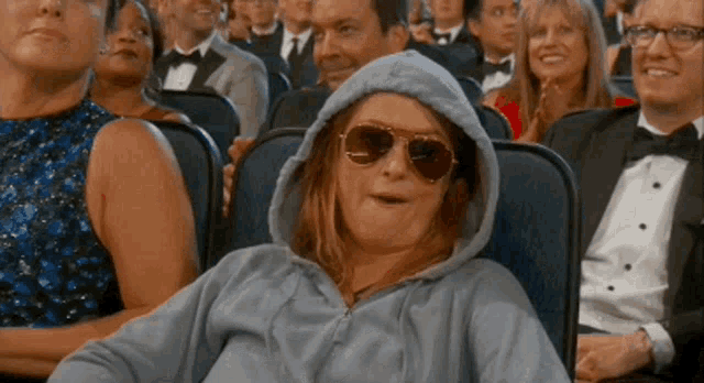 a woman wearing a hoodie and sunglasses is sitting in a theater .