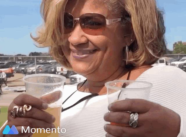 a woman wearing sunglasses and rings is holding a cup of beer and smiling