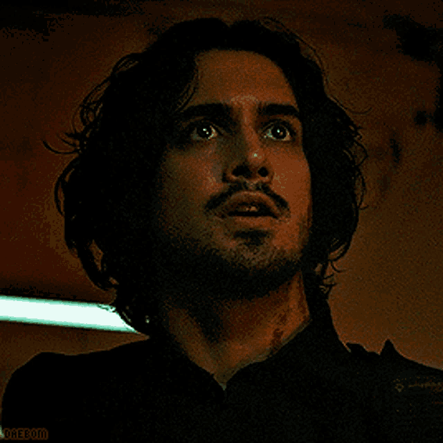 a man with long hair and a beard is looking up at something in the dark .