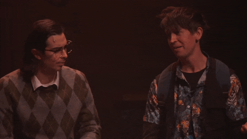 a man wearing glasses and a plaid sweater stands next to another man wearing a blue shirt with a tiger on it
