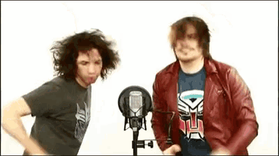 two men are standing next to each other in front of microphones . one has a transformer shirt on .