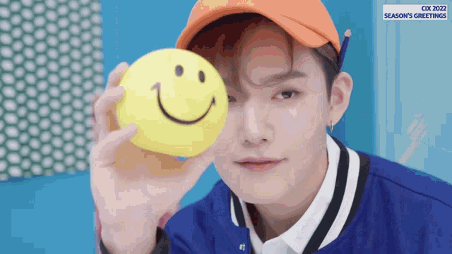 a man wearing an orange hat holds a yellow smiley face ball in front of his face