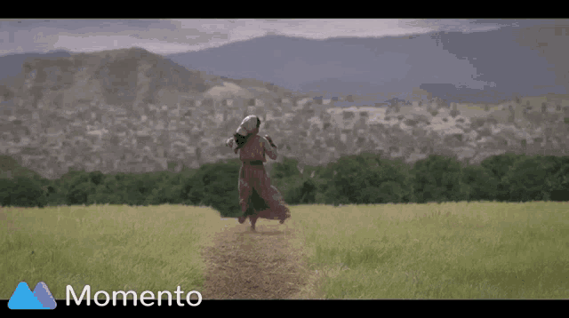 a woman in a dress is running through a grassy field with mountains in the background and the word momento in the corner