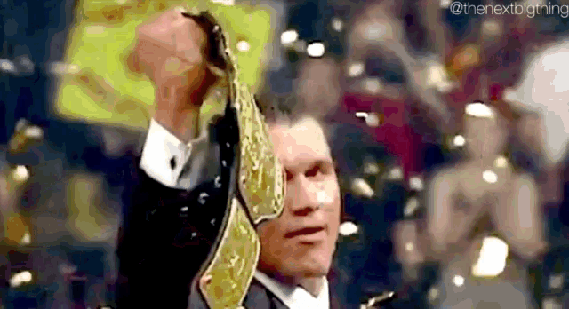 a man in a suit is holding a wrestling championship belt in his hand in front of a crowd .