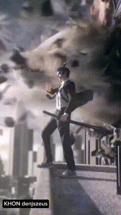 a man in a suit is standing on a ledge with the name khon denjszeus on the bottom right