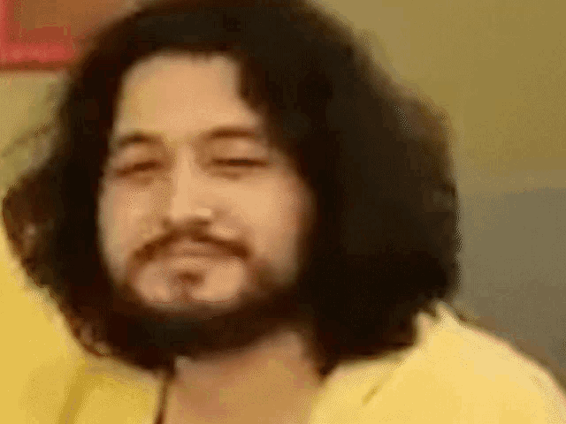a man with long hair and a beard is wearing a yellow shirt and making a funny face .