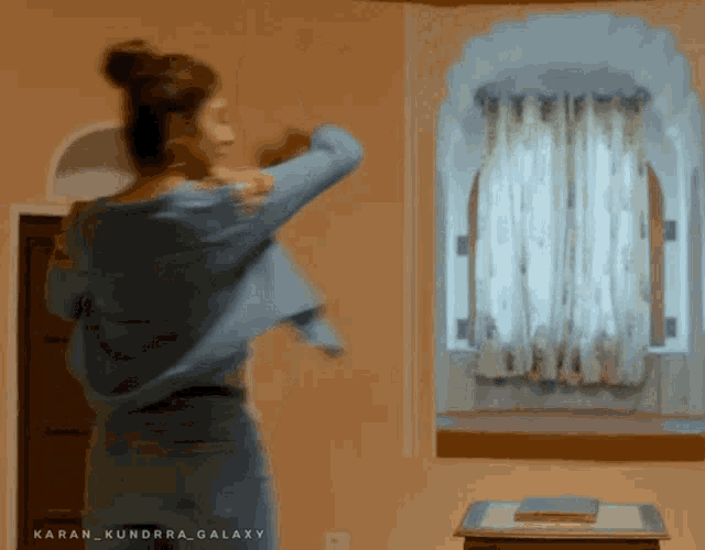 a woman in a blue jacket is standing in front of a window in a room with the words karan kundra galaxy on the bottom