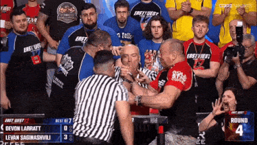a referee stands between two wrestlers in front of a crowd wearing east west jerseys