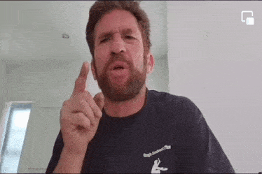 a man with a beard is wearing a black t-shirt and pointing his finger at something .