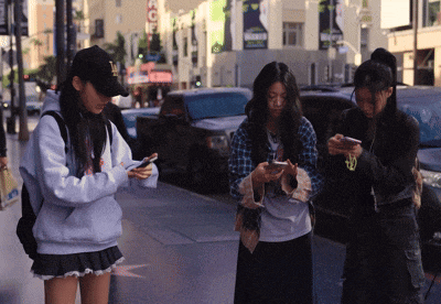 three girls are looking at their cell phones on a sidewalk in front of a building that says hollywood