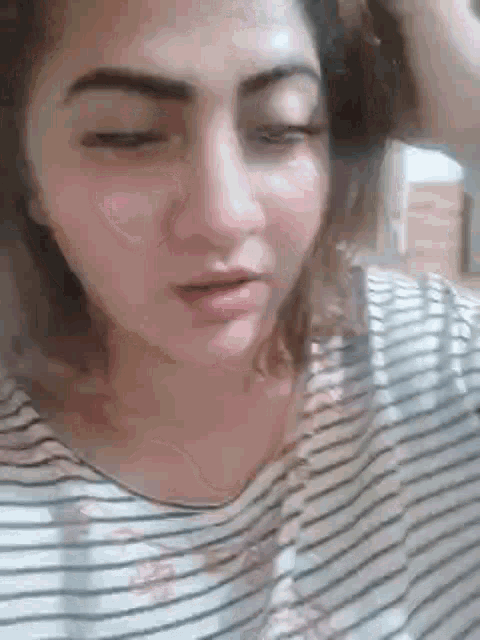 a woman in a striped shirt is looking at the camera with her eyes closed and her hand on her head .