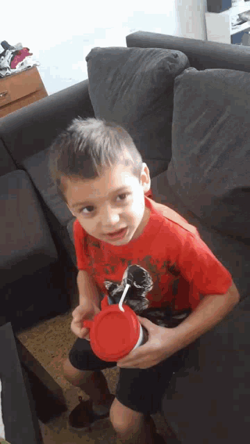 a young boy in a red shirt is sitting on a couch holding a cup with a straw