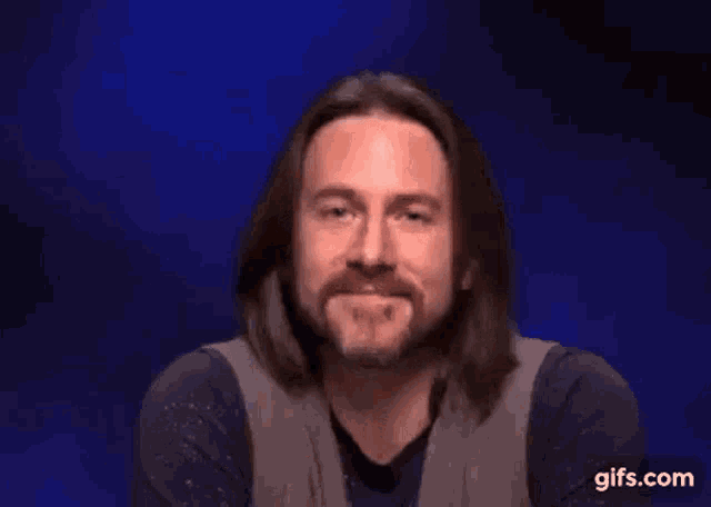 a man with long hair and a beard is waving his hand .