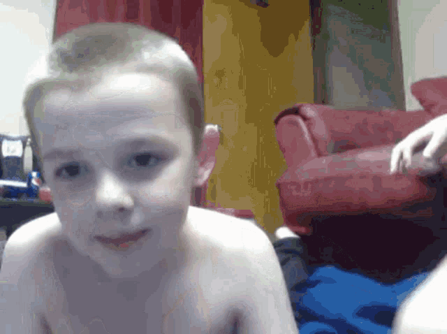 a young boy without a shirt looks at the camera with a red chair in the background