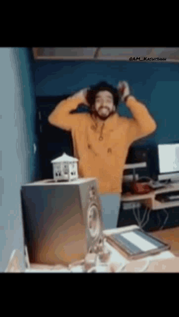 a man in a yellow sweater is standing in front of a computer and speakers