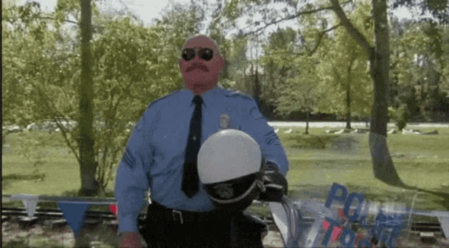 a bald police officer wearing sunglasses and holding a helmet stands in a park