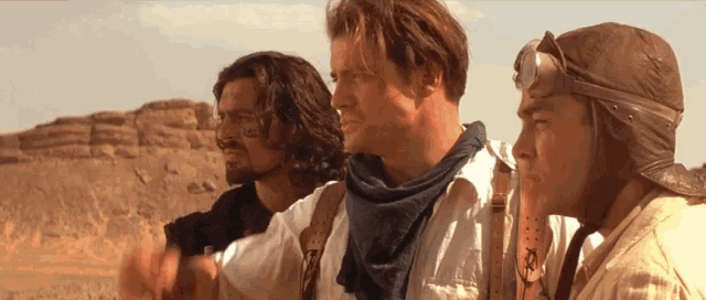 three men are standing in the desert and one has a helmet on his head