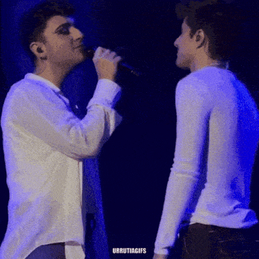 two men are standing next to each other and one is singing into a microphone while the other looks on ..