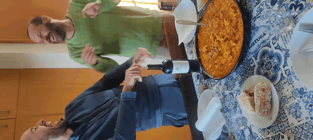two men are sitting at a table with a bottle of wine and a pan of food with a sign that says " ortiga "