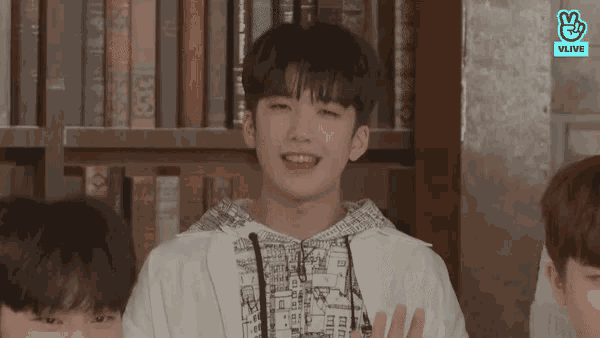 a boy in a white jacket stands in front of a bookshelf with a vlive logo on the bottom right
