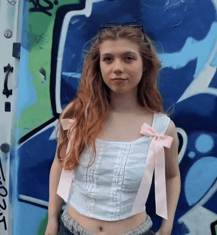 a woman wearing a white top with pink bows is standing in front of a wall with graffiti on it