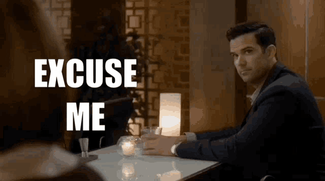 a man in a suit is sitting at a table with a woman and says excuse me .