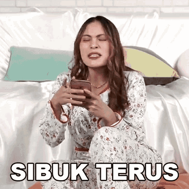 a woman is sitting on a bed holding a cell phone and the word sibuk terus is written above her