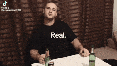 a man is sitting at a table with two bottles of beer and says " real " on the screen