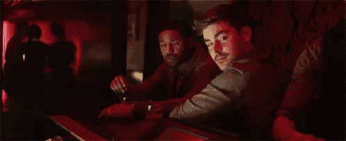 two men are sitting at a bar in a dark room in a red light .